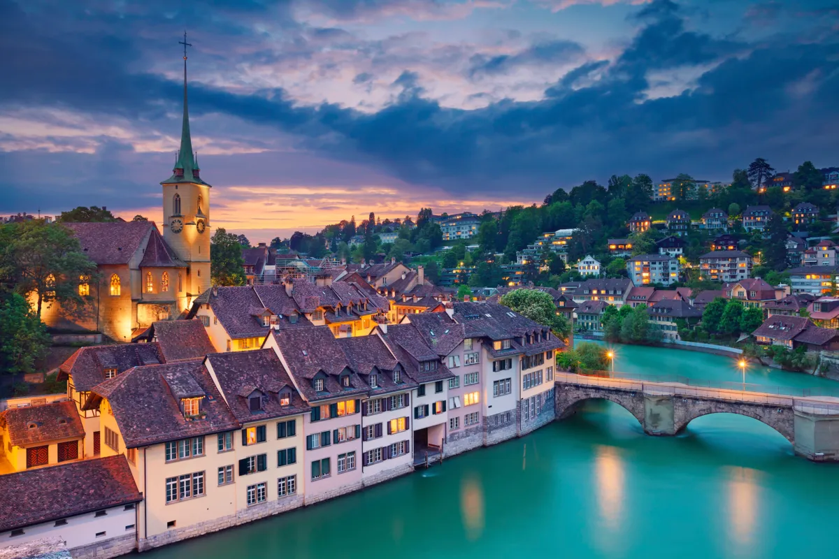 Panoramablick auf die malerische Stadt Bern: Die mittelalterliche Altstadt mit ihren historischen Gebäuden und den charakteristischen roten Ziegeldächern. Die imposante Zytglogge-Uhr und das majestätische Bundeshaus ragen stolz empor. Die Aare schlängelt